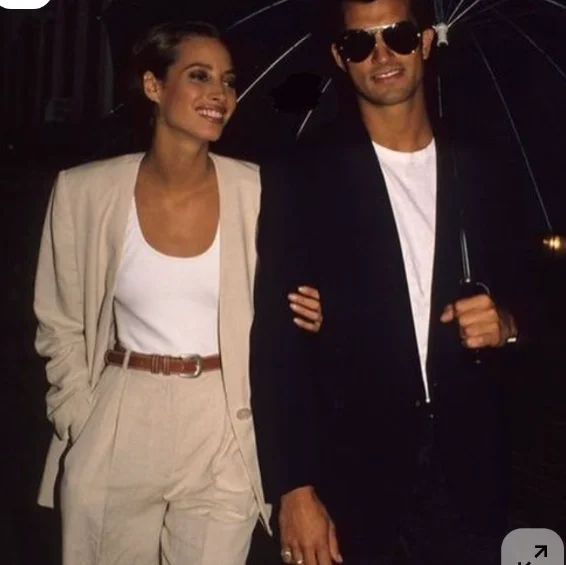 Un couple marche sous un parapluie, la nuit : elle en tailleur beige et débardeur blanc, lui en veste noire et lunettes de soleil.