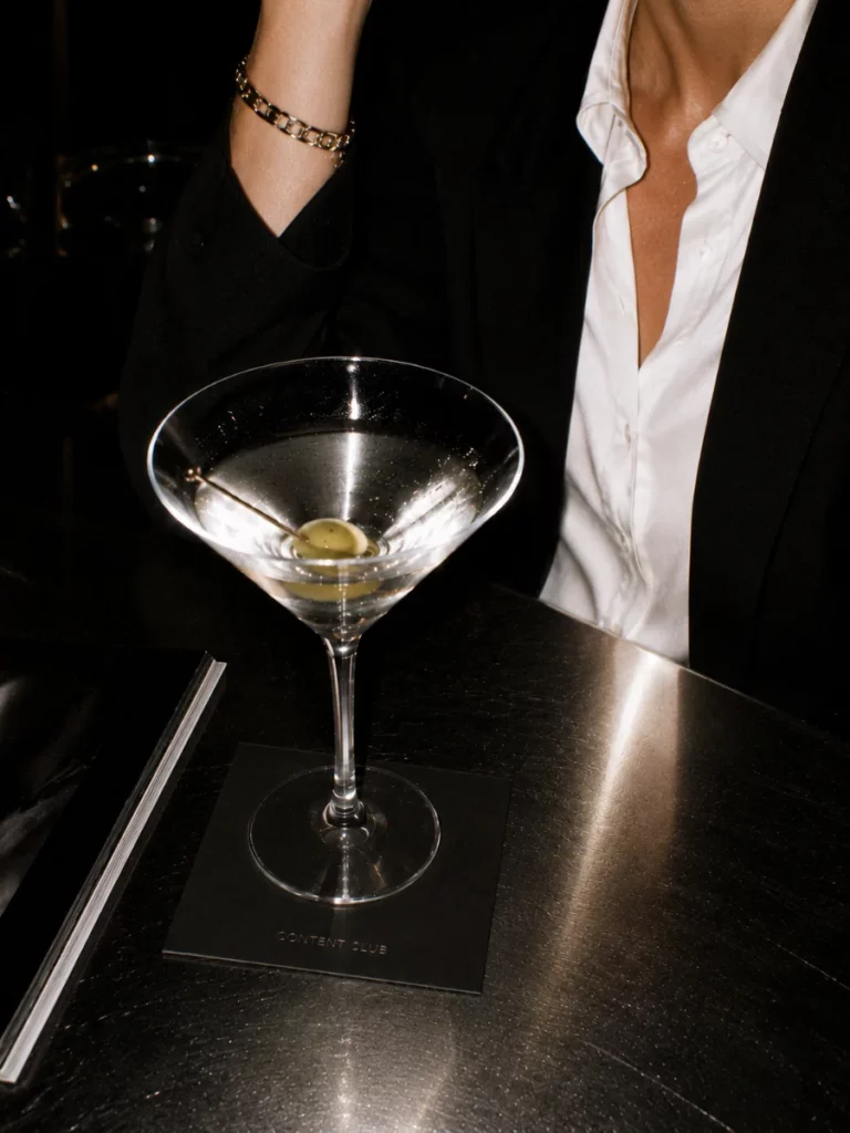 Sur le comptoir sombre d'un bar, un verre de martini garni d'une olive posé sur un sous-bock « Content Club » ; derrière, deux personnes en veste noire et chemise blanche ouverte.